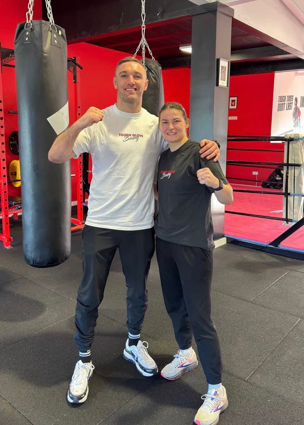 Tony Browne, in white, posing in a guard with a woman in a Tough Glove Boxing T-shirt beside the heavy bags