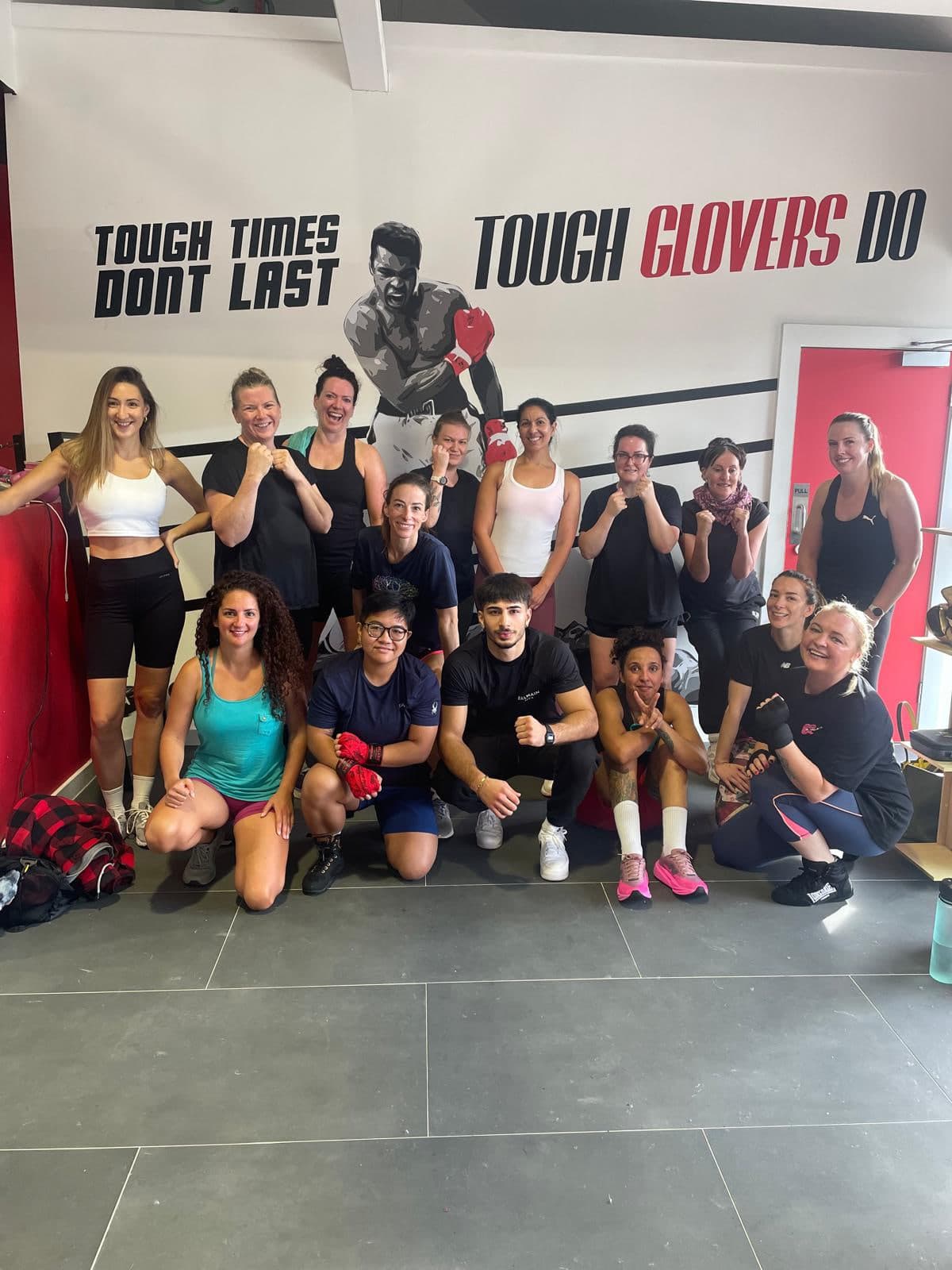 Members of a Tough Glove ladies class smiling together in front of the gym mural
